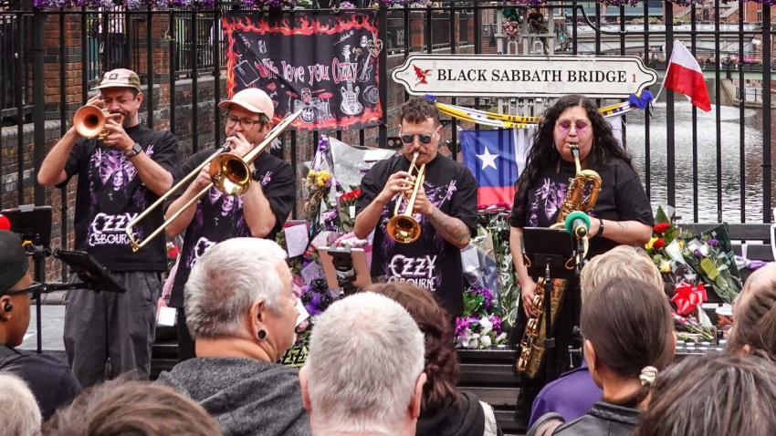 3F37YF0 Broad Street, Birmingham, 22 July 2026 - Fans of Ozzy Osbourne watched on as the band, Bostin' Brass played Black Sabbath songs in front of the Black Sabbath Bridge on Broad Street during Ozzy Day. Wednesday 22nd July is the one-year anniversary of Ozzy's death on July 22 2025. The city has put on various events and photo opportunities including having a Polaroid with the New Street Ozzy Bull that has been dressed in Ozzy's glasses, the inflatable giant Ozzy figure that was on display at the Back to the Beginning gig and visiting the Black Sabbath Bridge on Broad Street to lay tributes and pos